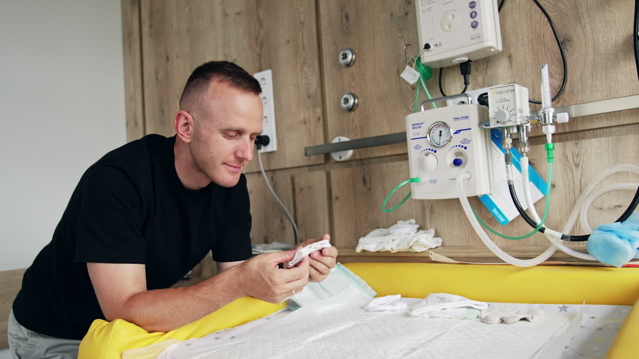 Caucasian mid-aged man wearing black t-shirt stands in the hospital ward. Expecting dad stacking up baby clothes.