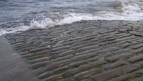 small waves of dirty sea water crashing on a concrete cobbled boat ramp in small fishing town in english seaside on grey day in slow motion