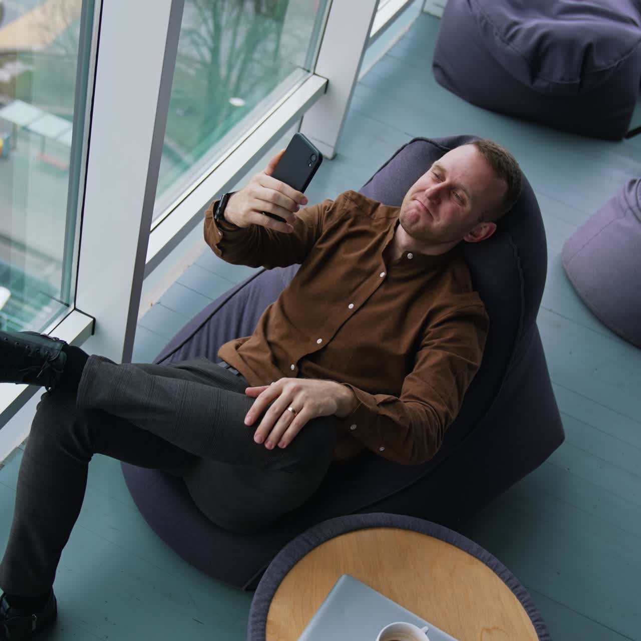 Businessman is having a video call. Young man in brown shirt and trousers sitting in a chair and talking online through the phone near the window. Top view