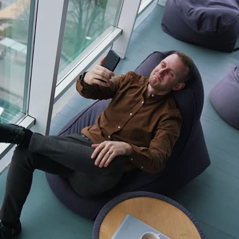 businessman is having a video call. young man in brown shirt and trousers sitting in a chair and talking online through the phone near the window. top view