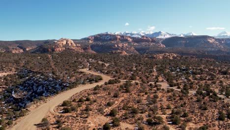 A-high-flying-drone-shot-over-a-remote-dirt-road-cutting-through-the-vast-and-unique-desert-land-near-Moab,-Utah,-with-the-snowy-Rocky-Mountains-towering-in-the-distance