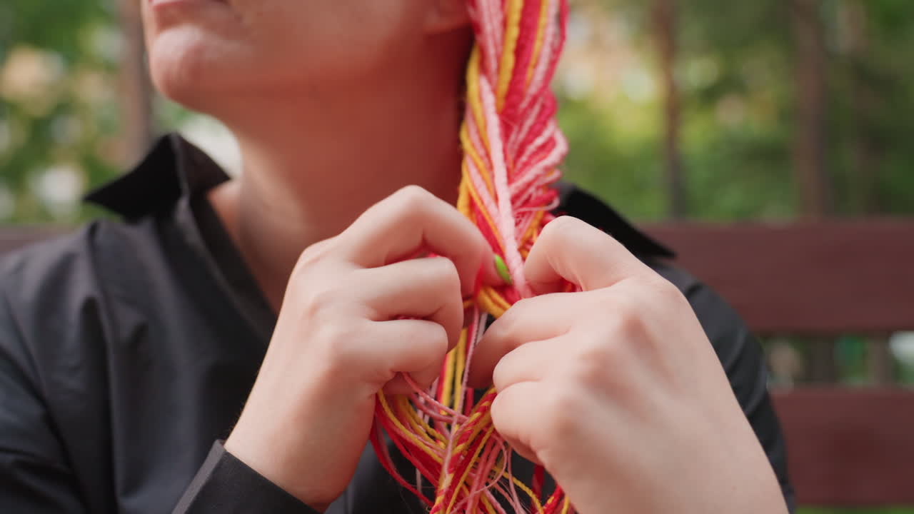 하얀 피부의 여성이 벤치에 앉아 알록달록한 땋은 머리를 정갈하게 땋고 있어요. 숙련된 손놀림과 차분한 움직임으로 머리카락 가닥과 텍스처에 집중하며 야외에서 머리를 손질하는 모습입니다.