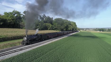 aerial view of an antique restored steam engine blowing smoke with passenger cars pulling out of a station on a sunny summer day