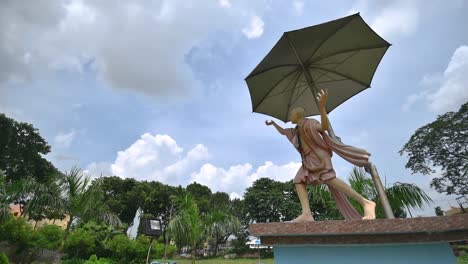 a timelapse of statue of indian demi god chaitanya deb with umbrella and clouds in blue sky near a hindu temple