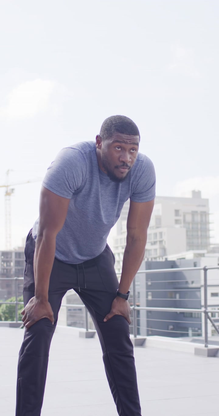 Vertical video: Man reaching and gripping 5kg kettlebell on roof in blue tee, gasping after set