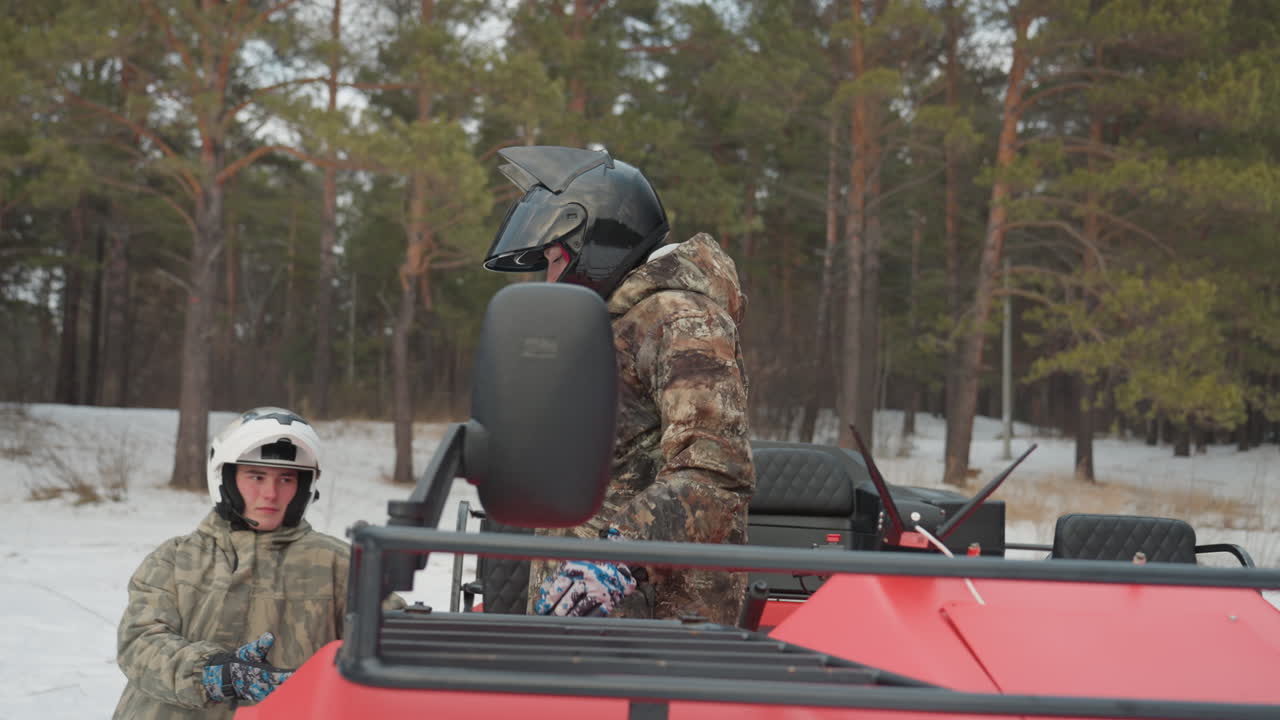 Controllo dell'equipaggiamento per il pilota di ATV in una foresta innevata. Un individuo vestito di nero prepara l'ATV tra gli alberi ricoperti di neve. Un caucasico, con abbigliamento mimetico, procede alla verifica del quad in un ambiente boschivo invernale.