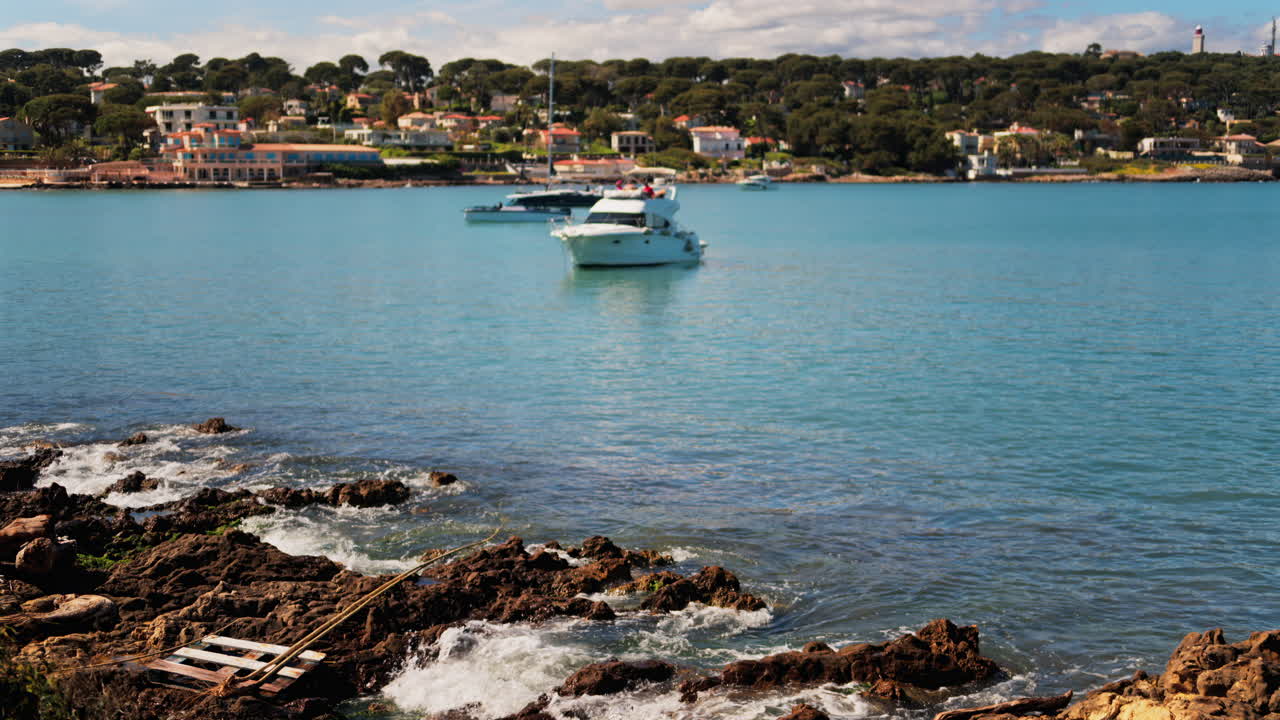 Waves crashing on the rocks on the shore with a boat floating on the sea and the town of Bandol, France on the background