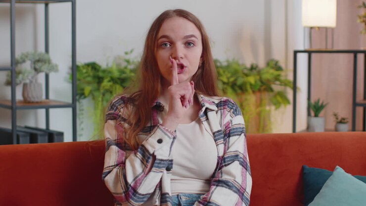 Young woman presses index finger to lips makes silence gesture sign do not tells secret at home
