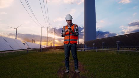 full body of asian male engineer with safety helmet using smartphone and looking around while standing with solar panel and wind turbines