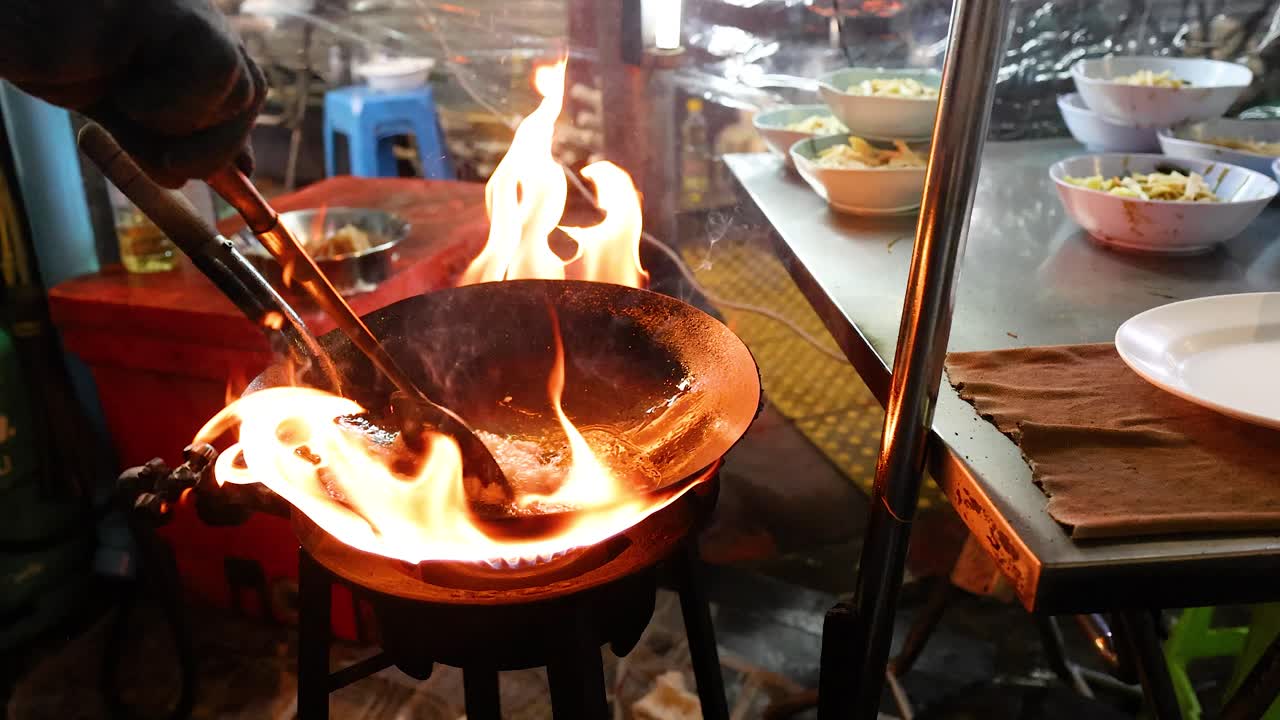 Chef stir-fries ingredients in flaming wok at night street food stall, dynamic lighting, handheld camera