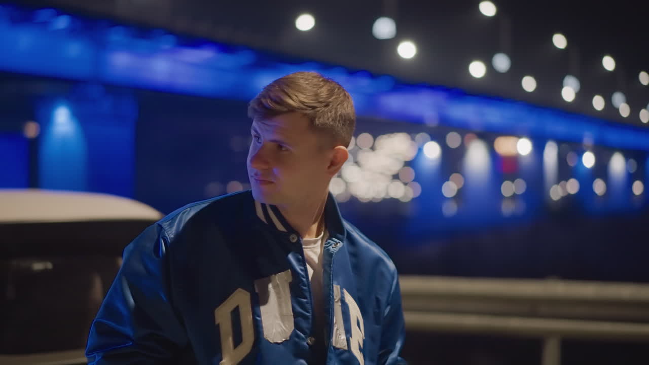 Night bridge portrait white driver near car, blue illuminated bridge lights and bokeh, contemplative young man in varsity jacket, waterfront reflections and city glow, cinematic urban atmosphere