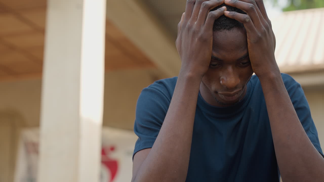 Distressed Black carpenter holding head outdoors at worksite, porch columns and roofline in background, visible sweat and tense posture, moment of worry over project setback, emotional candid portrait