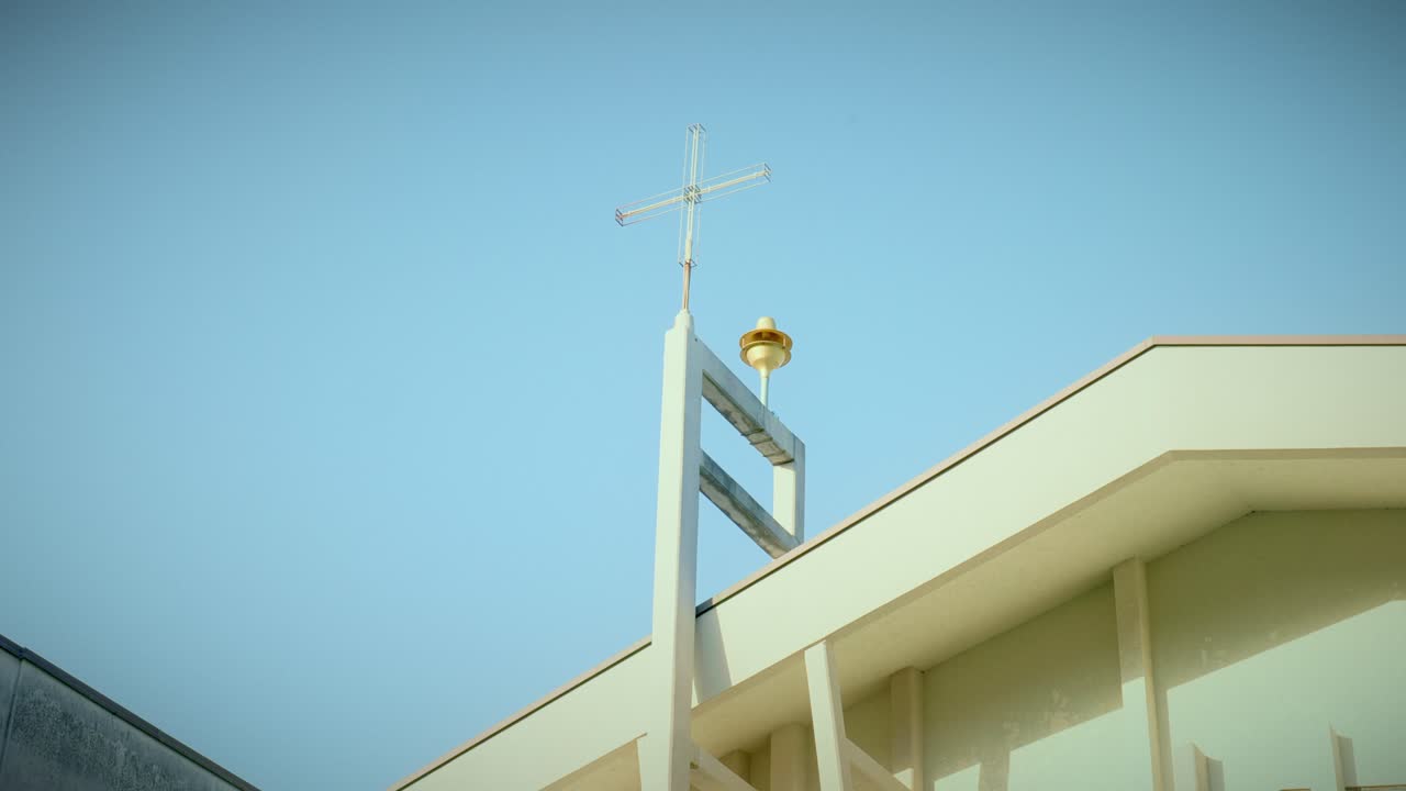 Ângulo baixo com inclinação ascendente da fachada de um prédio de igreja moderna com cruz fina e sino contra o céu azul claro na luz da manhã em Rimini