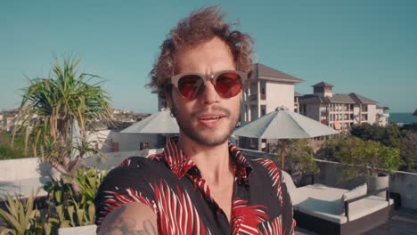 young man in sunglasses waving hands to the camera in the foreground, terrace with pool and hammocks in the background