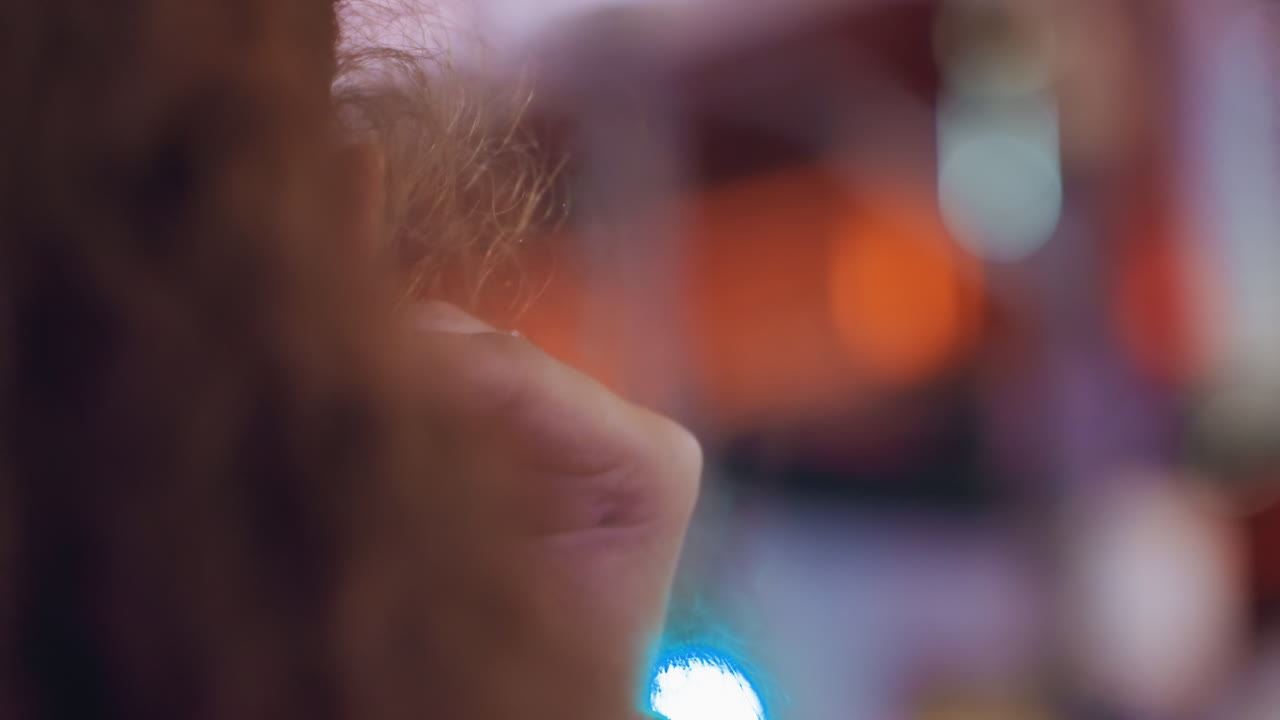 Nahaufnahme im Profil: Eine Person richtet unter Neonlichtern einen Ohrring, Haare im Dutt, gemusterte Brille, kleine Creolen-Piercings und aufmerksame Geste. Eine authentische urbane Nachtszene mit farbigem Bokeh und intimen Details.
