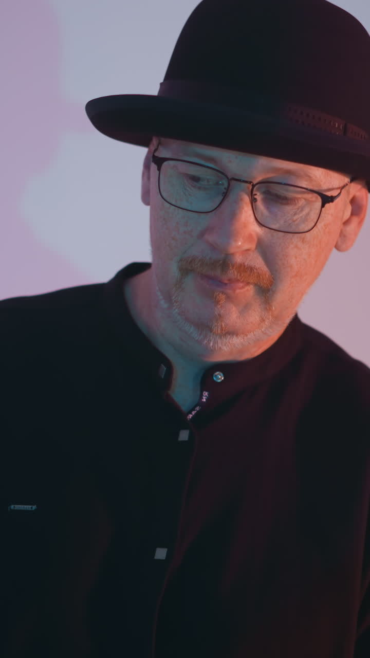 Close-up view of a man in a black shirt, black hat, and glasses standing against a white background with a soft gradient of blue and pink light, creating a thoughtful and introspective mood