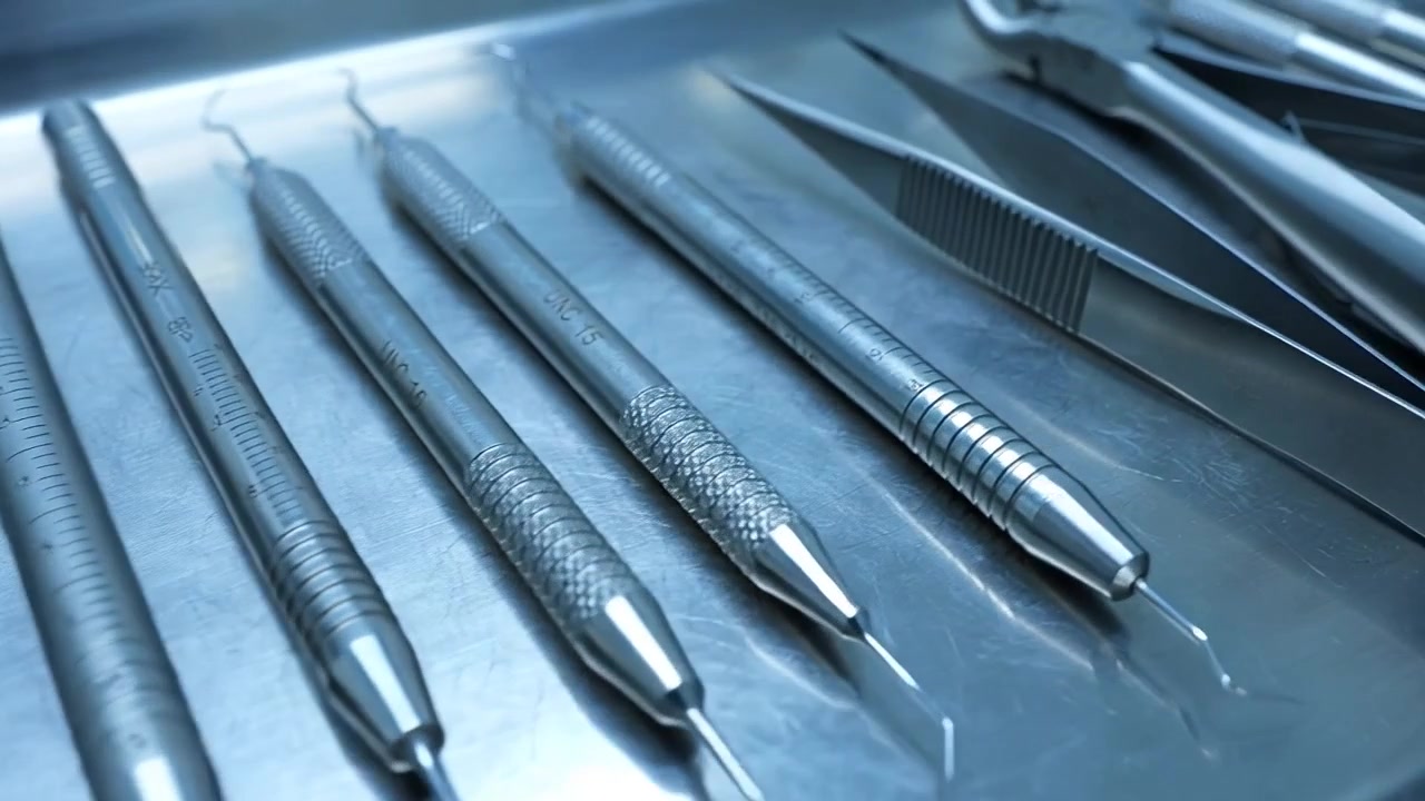 Stainless steel dental instruments including mirror, probe and tweezers arranged on a metal tray under cool light