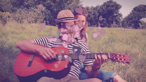 animation of hearts floating over caucasian couple sitting on grass and playing guitar