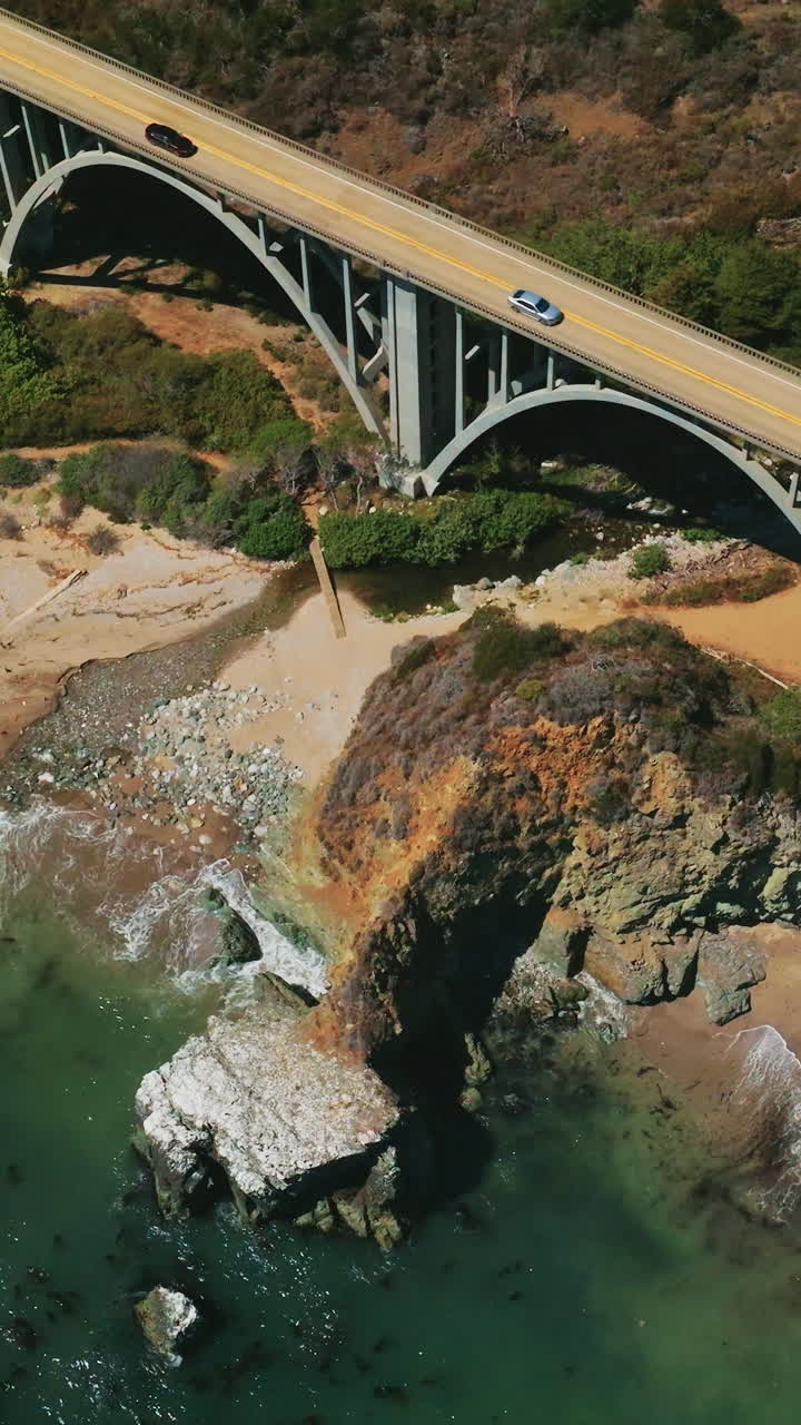 Beautiful arch bridge close to the sandy beach in California mountains. Blue water splashing waves on the shore. Aerial view. Vertical video
