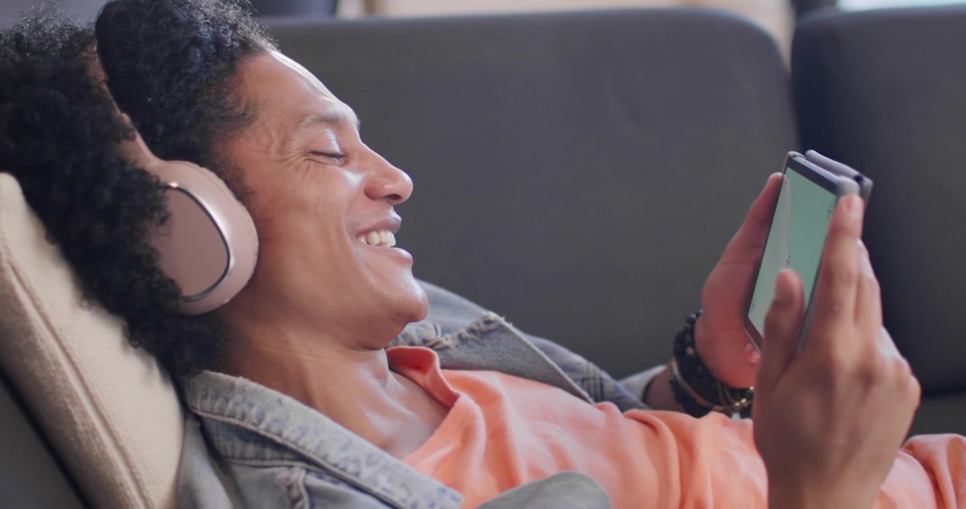 Uomo afroamericano disteso sul divano dopo una notifica, con le cuffie, che tocca il telefono e ride.