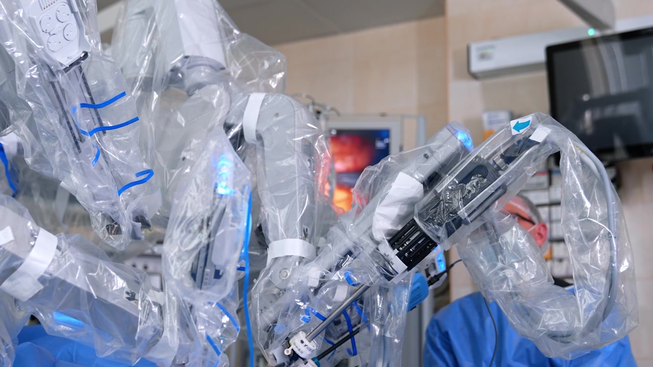 Surgical robotic system moves operating the patient. Male surgeon sits at backdrop focused on the monitor.