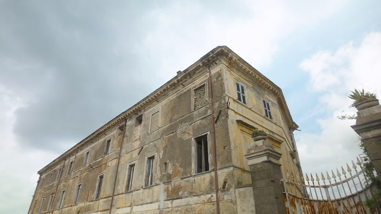 Aged building in Castel Gandolfo with worn facade and broken windows beneath a cloudy sky.