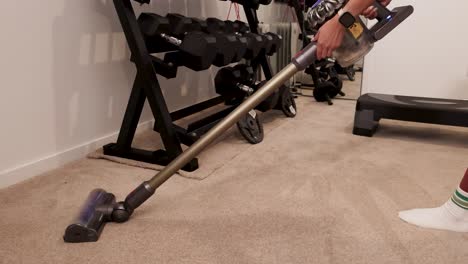 a person vacuums a carpeted home gym with weights, using a cordless vacuum cleaner. bright lighting enhances the clean, organized space