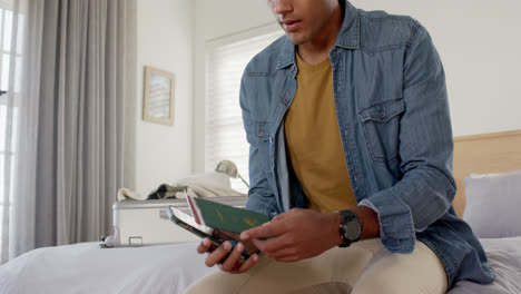 hombre biracial usando un teléfono inteligente y comprobando el boleto y el pasaporte en un dormitorio brillante