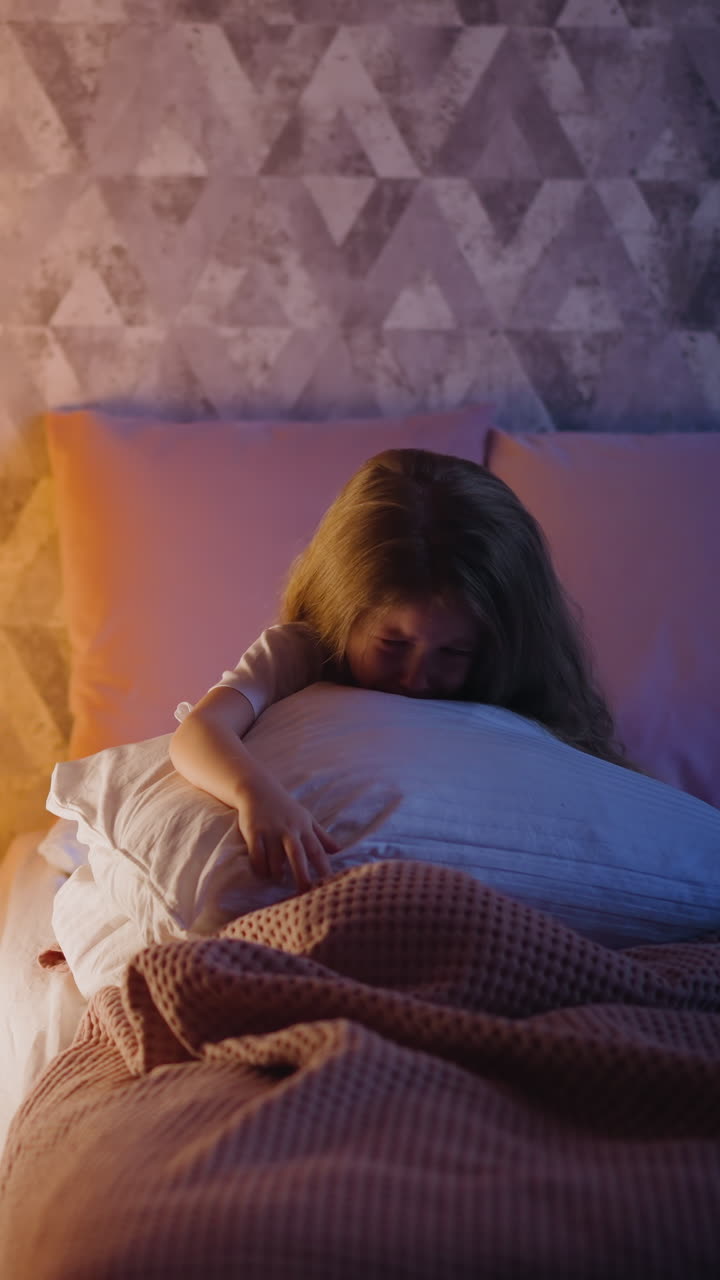 Sad little child hugging huge pillow cries sitting on bed in semidark room partially illuminated by night lamp glowing inside play teepee in evening slow motion