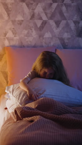 sad little child hugging huge pillow cries sitting on bed in semidark room partially illuminated by night lamp glowing inside play teepee in evening slow motion