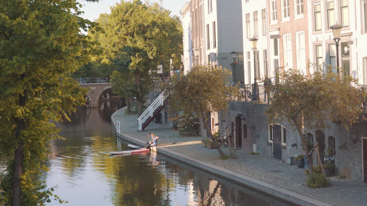 Two Ladies Sitting By The Riverbank Of Oudegracht Canal In Utrecht, Netherlands On A Sunny Morning - drone shot (backward)