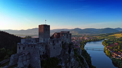 flag of slovakia waves in the wind on the top of the strecno castle. beautiful river crossing the picturesque village at backdrop