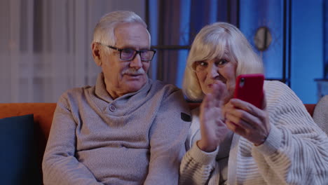 Sonriente-Pareja-De-Ancianos-Abuela-Abuelo-Haciendo-Videollamadas-En-Línea-En-El-Teléfono-Móvil-En-El-Sofá-De-Casa