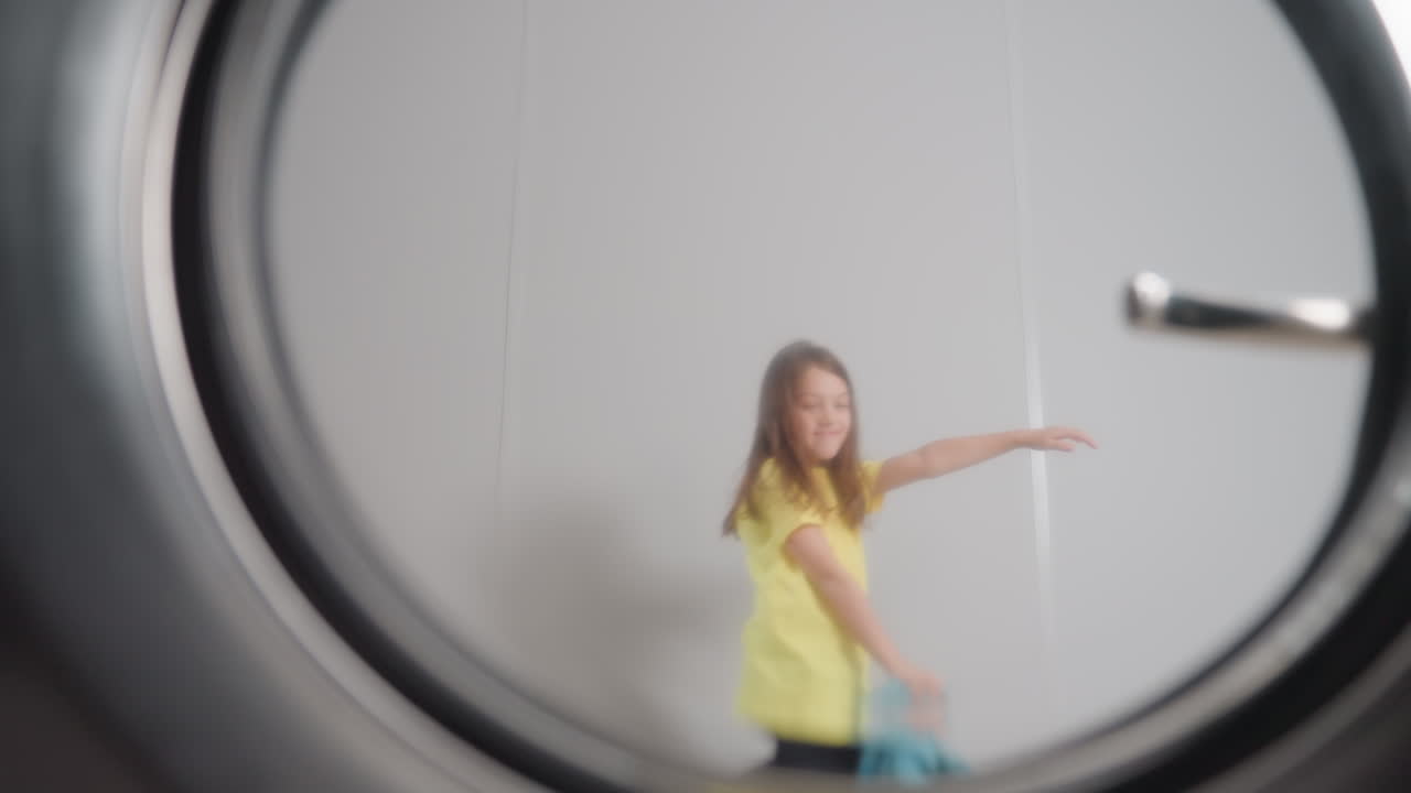 Washer door window shows kid dancing past in soft reflection, round handle visible at right, motion blur around frame, bright interior, joyful laundry moment, playful energy, pov from drum rim