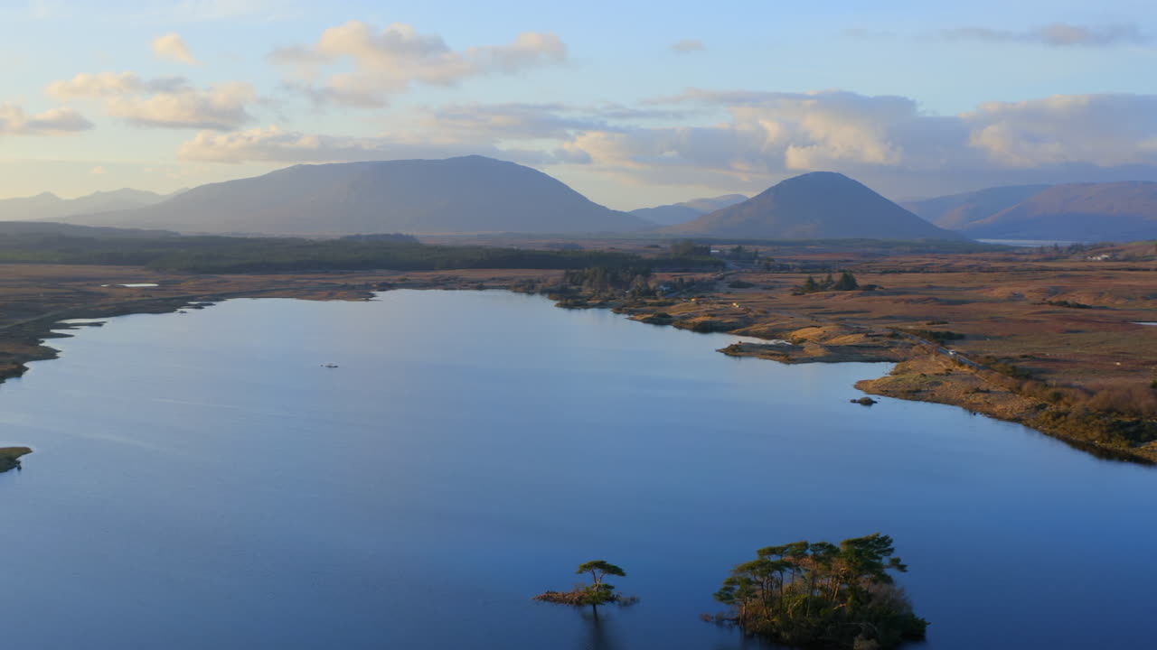 Aerial orbit over Lough Bofin at golden hour, a warm glow over the tranquil waters and surrounding rolling hills, Connemara, Galway, Ireland