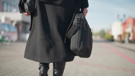 close-up of a woman walking confidently in a chic black coat and boots, carrying two bags, one hand adjusting her coat on a sunlit urban street and blur view of parked bike