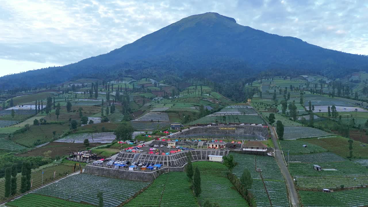 Aerial view of a camping area filled with tents on a mountain slope surrounded by green plantation land. Embung Bansari, Mount Sindoro, Indonesia.