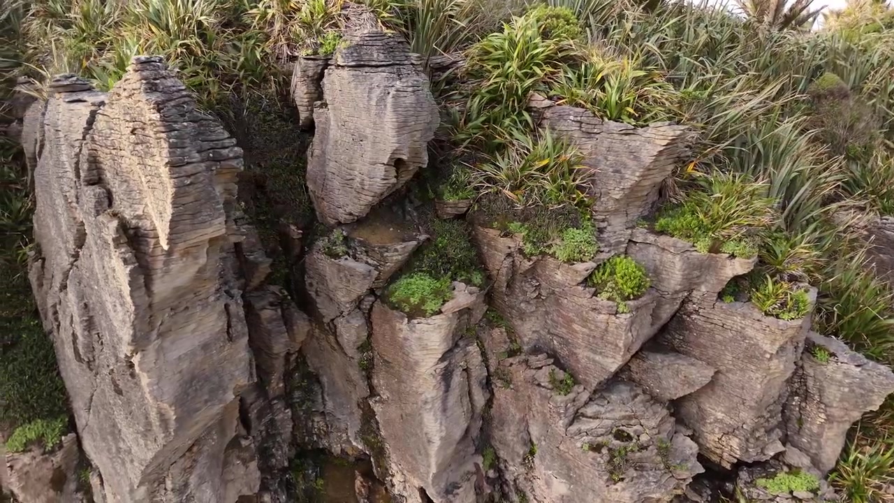 Drone panoramaoptagelse glider fra vidvinkel til nærbillede af Pancake Rocks ved solnedgang i Punakaiki, New Zealands vestkyst. Bølger slår mod detaljerede lagdelte kalkstensformationer under gyldne himmelstrøg