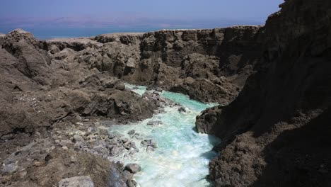 Drone-shot-Flying-low-through-a-rugged-steep-desert-canyon-in-wilderness-over-fast-running-crystal-clear-hot-spring-waters-to-the-Dead-Sea-on-a-sunny-day-with-blue-sky-in-Israel