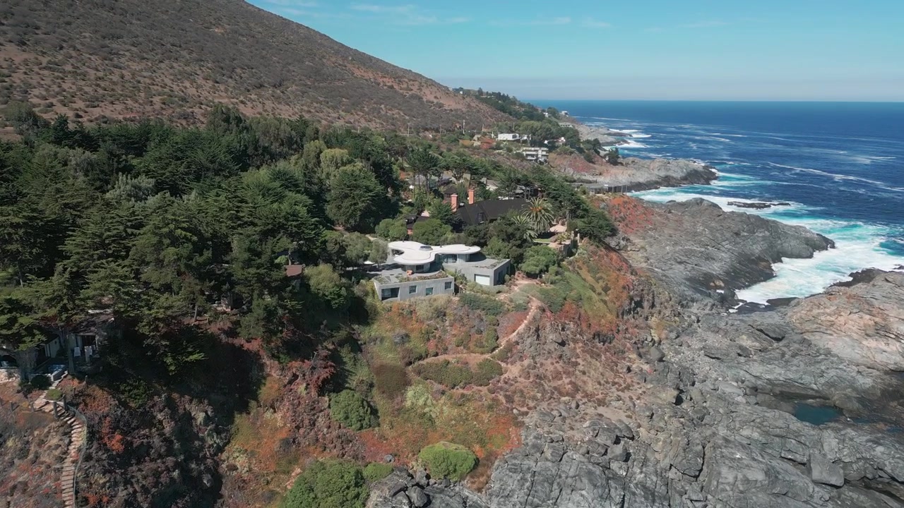 Vuelo rasante sobre un acantilado escarpado con villas modernas integradas en densa vegetación nativa sobre una espectacular costa rocosa con olas rompiendo. Punta Pite, Papudo, Chile