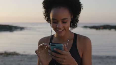 portrait of young african american woman wearing earphones listening to music using phone texting browsing on beautiful sunset beach