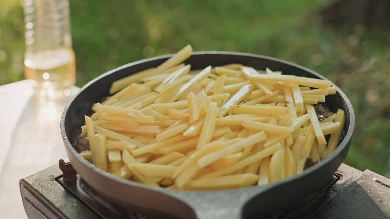 Skillet piled with raw fries cooking outdoors on portable camping stove, soft sunlight, calm backyard meadow, golden oil slowly warming, camper prepping simple comfort snack for relaxed morning