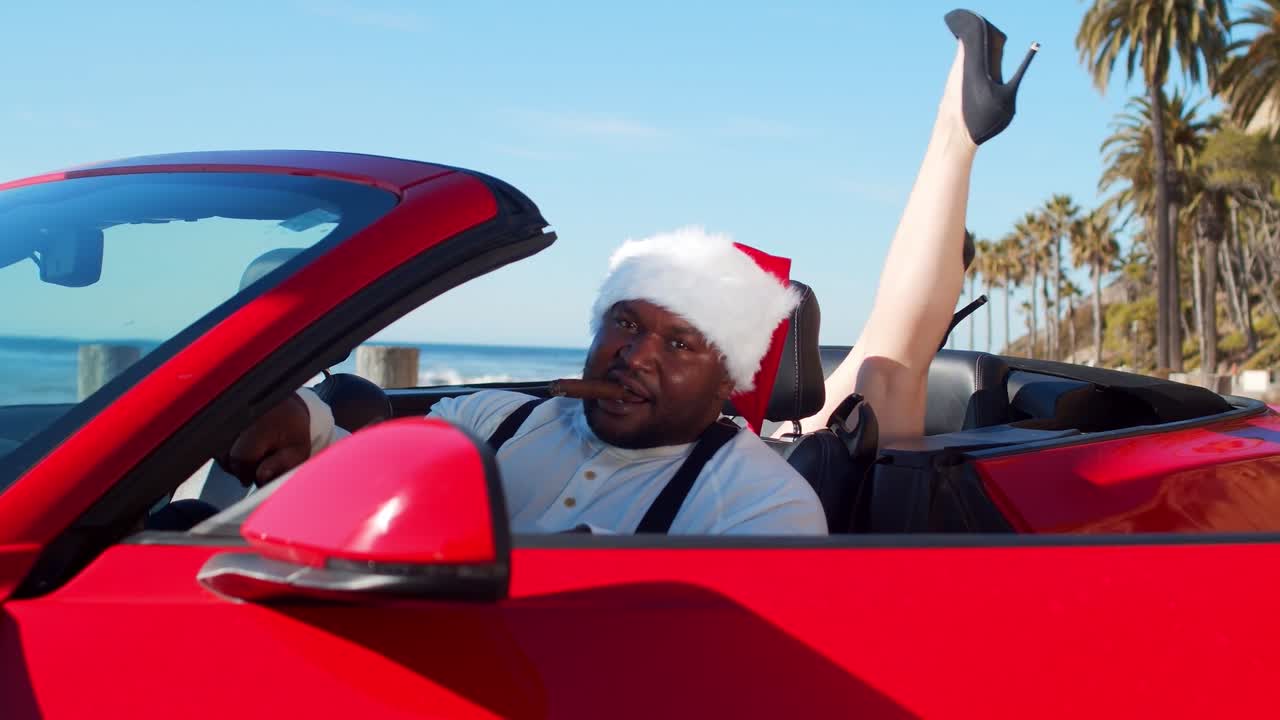 vídeo en cámara lenta de un hombre negro con un gorro de papá noel en un coche rojo en la playa con una mujer con tacones altos levantando los pies en la parte trasera del coche mientras graba un vídeo musical.
