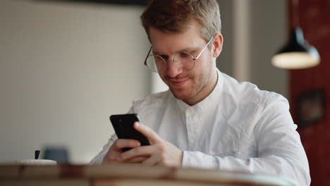 Un-Joven-Con-Gafas-Descansa-En-Una-Cafetería-Y-Charla-En-Línea-En-Las-Redes-Sociales-Mediante-Un-Teléfono-Móvil-Con-Wifi-Gratuito-E-Ilimitado