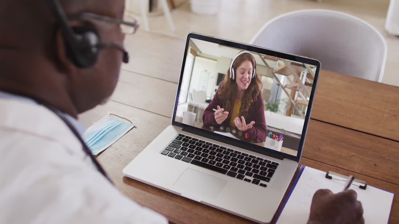 Afroamerikansk forretningsmand sidder ved skrivebord, bruger bærbar computer og har videoopkald med en kvindelig kollega.