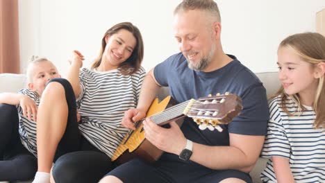 an adult man sings a song and plays the guitar for his family sitting on the couch at home