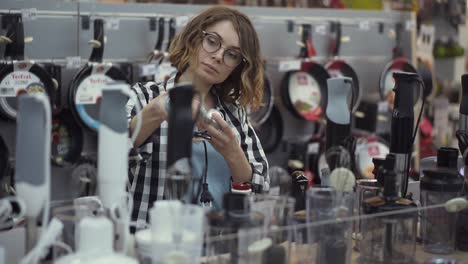 in the appliances store, an attractive curly woman in a plaid shirt chooses a blender stick by viewing and holding the device in her hands. slow motion