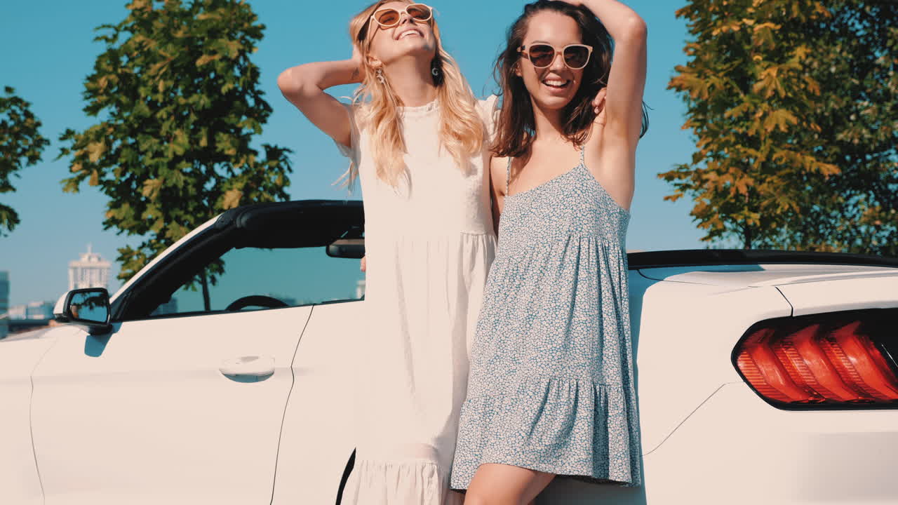 여름 드레스를 입은 두 명의 행복한 여성들이 색 컨버터블 자동차에 의해 포즈를 취하고 있습니다.