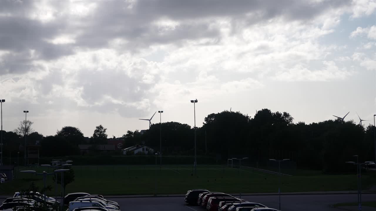 A wide establishing shot of Falkenberg’s soccer stadium parking lot, with a modern wind turbine spinning in the background under partly cloudy skies.