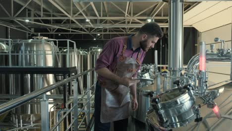 young male brewer wearing a leather apron supervise the process of beer fermentation at a modern brewery factory